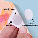 Hand holding white backing paper with circular hook fastener dot showing textured surface. Annotation text reads "Adhesive backs", "Hook dot", and "Loop Fasteners available separately". Colourful painted artwork visible in background on pale blue surface.