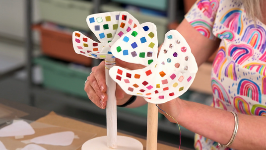 Hands holding two white Magiclay flower sculptures mounted on wooden dowels, each decorated with small, colourful mosaic-like squares and reflective embellishments. The flowers are shown upright on a work surface, mid-construction, in a studio setting.

Image credit: Zart Education On Demand: Magiclay Mosaic Flowers Inspired By Skunk Control, by Dee Zabel.