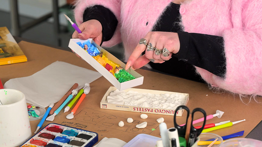 Miniature Holiday Diorama, a Zart Education On Demand lesson plan activity with Francesca Spillane. Hands in a pink faux-fur cardigan use a purple sculpting tool to shape a paper box scene with blue, yellow and green textured clay hills dotted with tiny figures, surrounded by modelling tools and paint.
