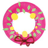 Zart Paper Plates crafted into a Christmas wreath, with a hot pink plate ring decorated with green paper leaves, yellow paper circle baubles, and small orange and purple pom-poms. Finished with a red and gold tinsel bow at the base, made from twisted glittery pipe cleaner-style trims.