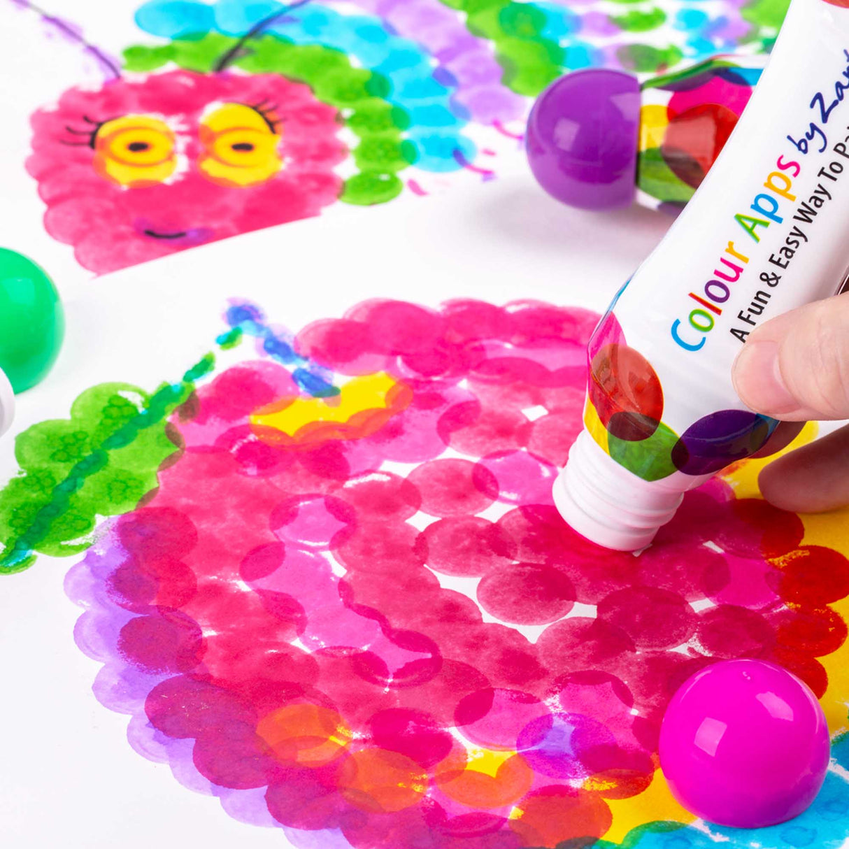 Zart Colour Apps bottle being pressed onto paper to create overlapping pink, purple and yellow circular marks. A bright pink cap sits nearby, showing how the rounded sponge tip creates layered dot textures.