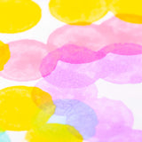 Close-up detail of overlapping pastel paint dots on white paper. Soft pink, yellow, purple and blue circular stamps blend translucently, showing layered colour mixing and the textured effect created by sponge tip application.