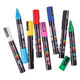 Assorted Uni POSCA paint markers scattered on a white background, including white, yellow, pink, red, blue, black and green. One blue marker is uncapped, revealing the bullet tip.