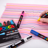 Hand using a yellow POSCA marker to draw curved coloured lines on pink paper for an optical-illusion style hand artwork. POSCA markers and packaging sit around the page.