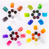 Four open round Micador Brilliant Water Colours discs are surrounded by painted swatches on white paper. Colours include greys, browns, greens, blues, yellows, oranges, reds, pinks and purples, with soft wash effects, feathered edges and varying pigment density.