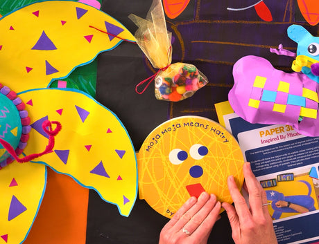 Hands holding a yellow circular artwork with text "Moja Moja Means Hairy" and a simple face design, surrounded by colourful paper craft projects including decorated yellow fish, a purple tortoise with geometric patterns, and hanging mobiles. Planning a Primary Art Program booklet visible showing "PAPER 3D Inspired By Mitah" lesson plan.