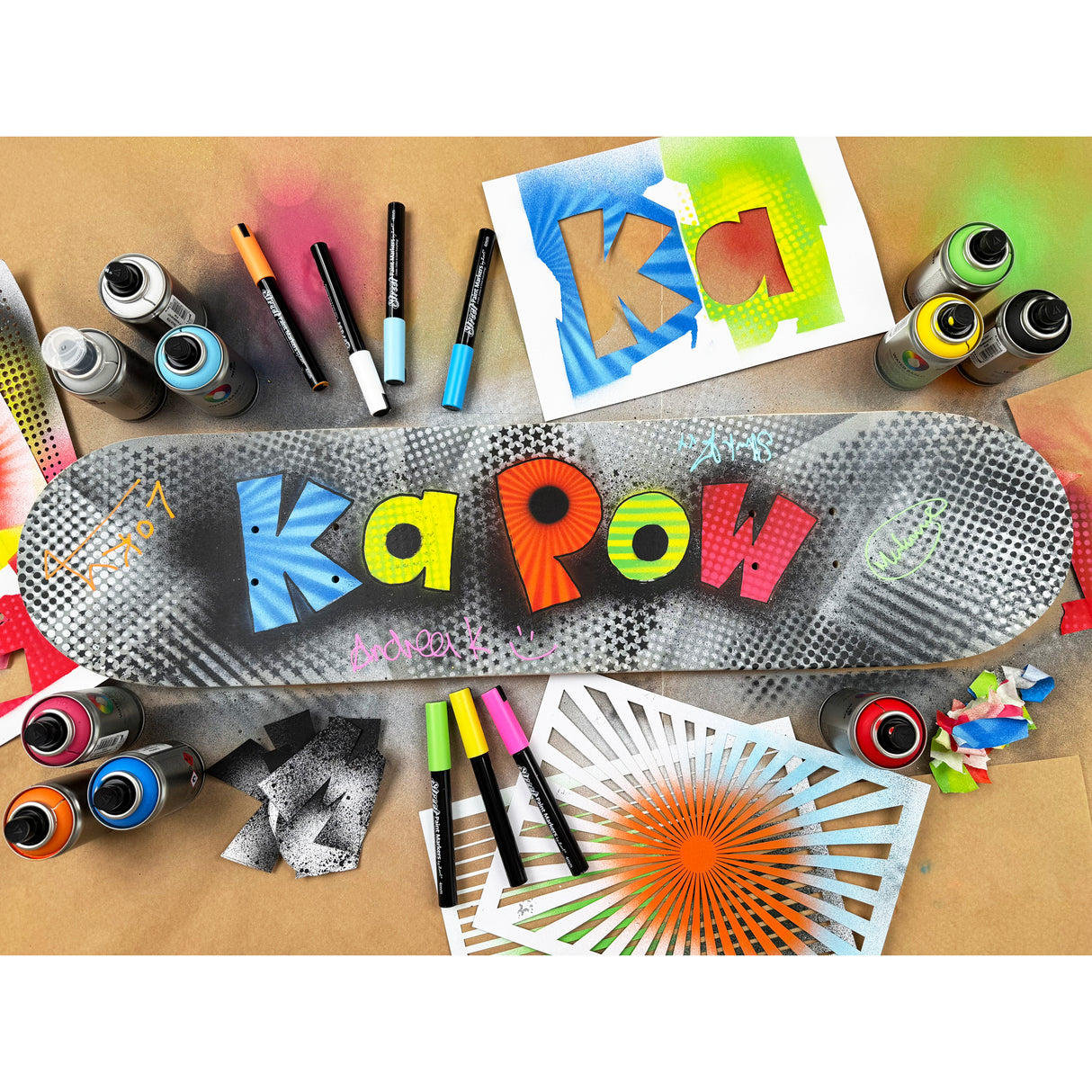 Decorated skateboard deck surrounded by spray paints, markers and stencil sheets. The deck features bold graffiti-style lettering reading “Ka Pow”, with handwritten tags layered over stencilled dots and textures.