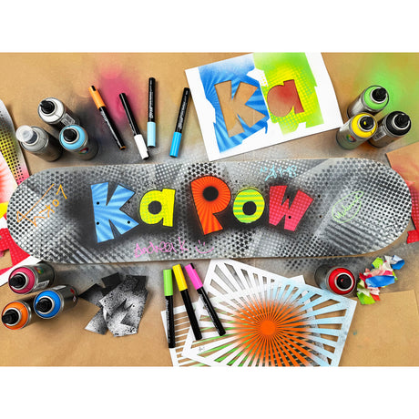 Decorated skateboard deck surrounded by spray paints, markers and stencil sheets. The deck features bold graffiti-style lettering reading “Ka Pow”, with handwritten tags layered over stencilled dots and textures.