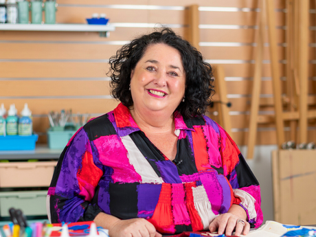 Zart Education PL Artist Facilitator Tania Di Berardino – A woman seated in an art classroom, smiling at the camera while surrounded by colourful craft materials including yarn, fabric pieces and bottles of paint. Shelving with brushes, paints and art tools is visible in the background, creating a vibrant and creative studio setting.